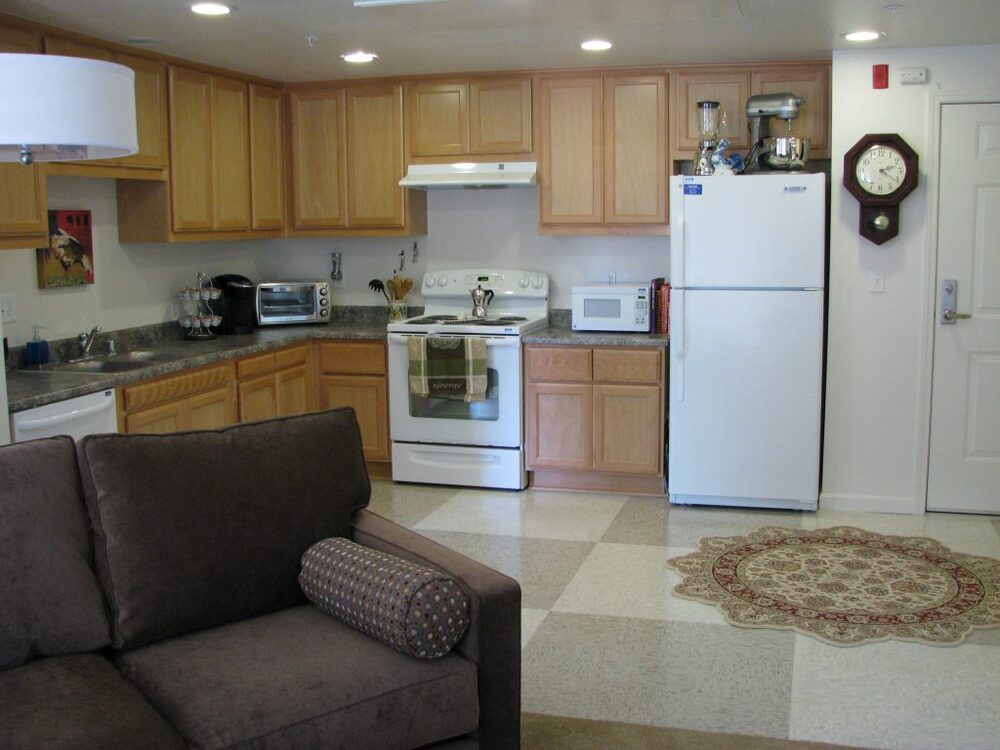 The corner of the couch is on the left and the kitchen opens up behind it. There are appliances on the counters and on top of the refrigerator. 