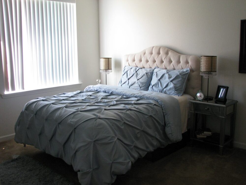 A bed with lamps on nightstands on either side of it. Sun light streams in through window blinds. 