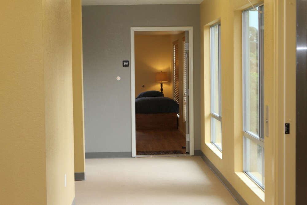 A clean hallways leads to an open room with a bed and a lamp in it. 