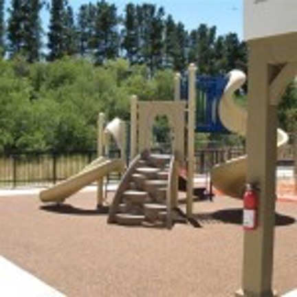 A kids playground with slides and stairs to climb sits  in an enclosed black fence. There are tall pine trees in the distance. 