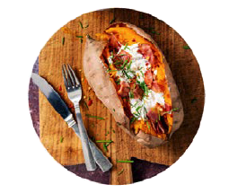 A fork and spoon sit on a wooden cutting board next to a baked sweet potato that is covered in toppings.